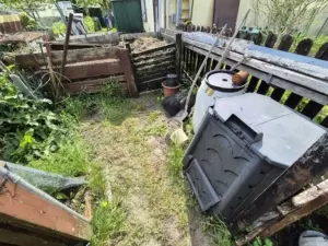 Zwei Holzkomposter in einem verwilderten Garten, umgeben von Unkraut und jungen Pflanzen.