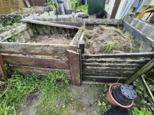 Zwei verwitterte Holzkomposter gefüllt mit Gartenabfällen und umgeben von Unkraut, in einem Gartenbereich.