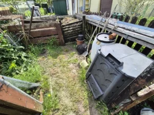 Zwei Holzkomposter in einem verwilderten Garten, umgeben von Unkraut und jungen Pflanzen.