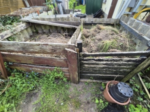 Zwei verwitterte Holzkomposter gefüllt mit Gartenabfällen und umgeben von Unkraut, in einem Gartenbereich.